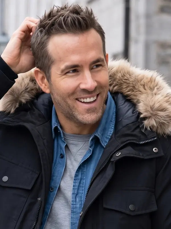 A smiling man wearing a black winter parka with a tan fur hood lining, layered over a blue denim shirt and a gray t-shirt, with his hand in his hair.
