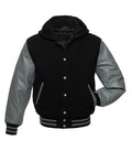 Front pose view of a black wool hooded varsity jacket with grey leather sleeves, silver snap buttons, and one hand inserted in the side pocket.